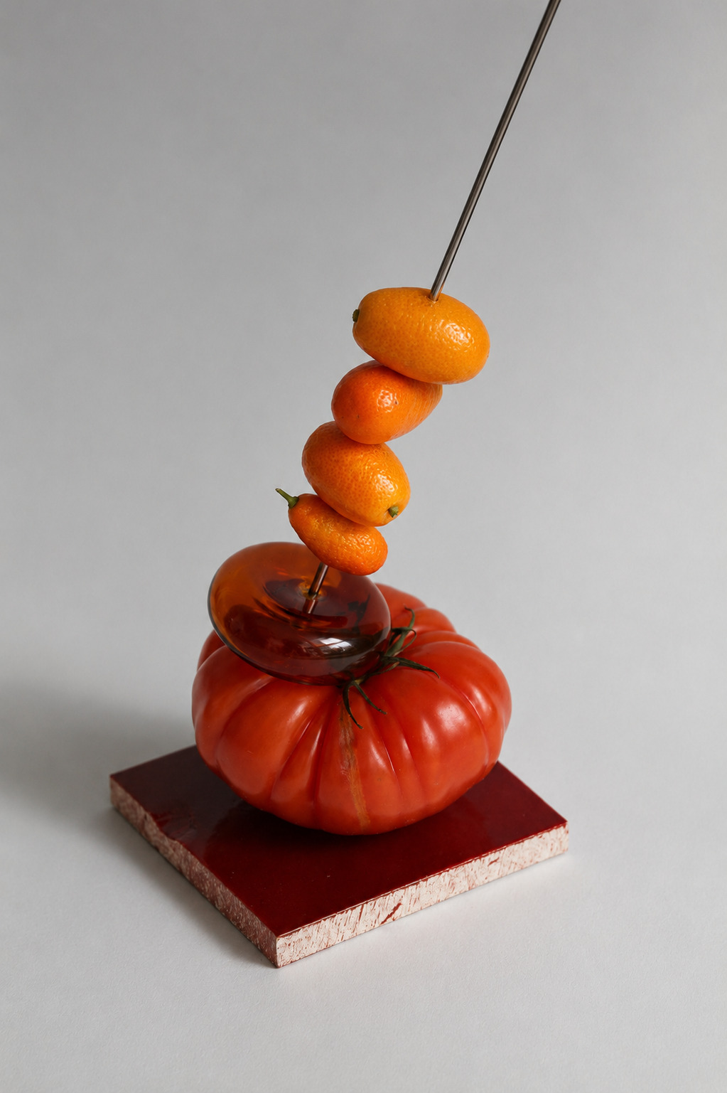 Sculptural still life of skewered kumquats balanced above a heirloom tomato